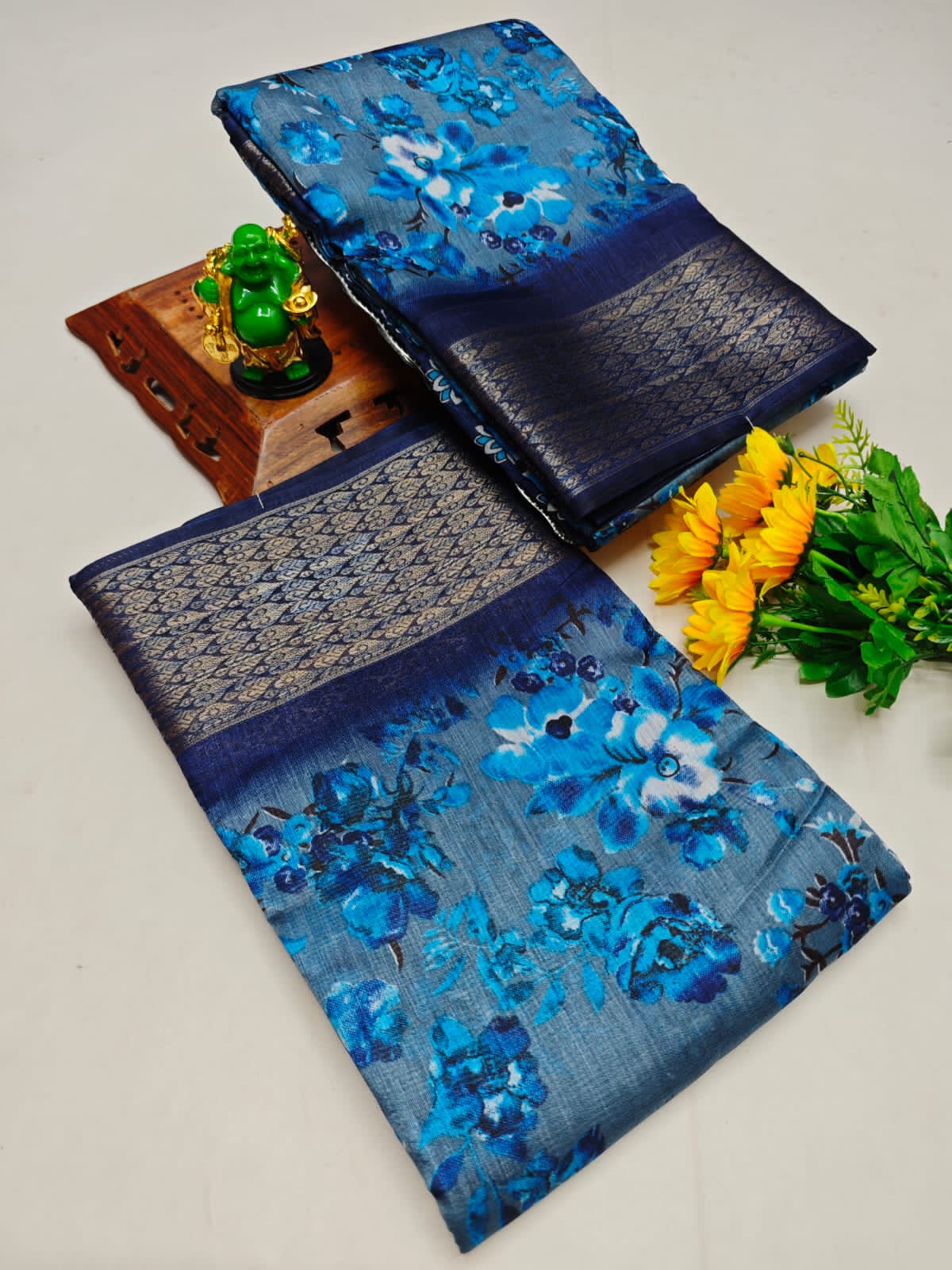 Two blue floral sarees with a small statue and flowers on a light gray background